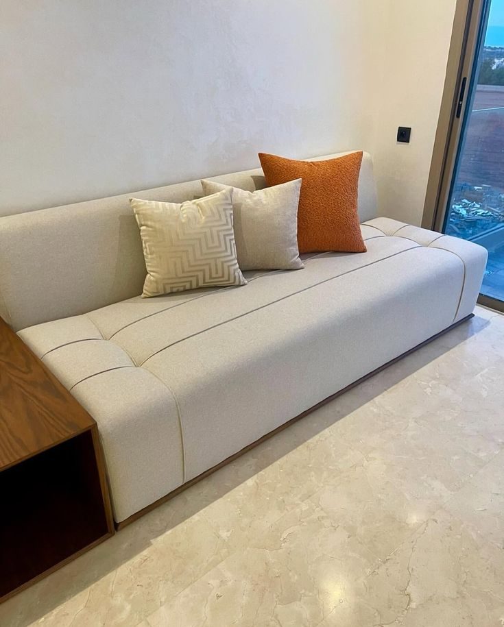 Banquette basse crème à coussins orange devant une baie